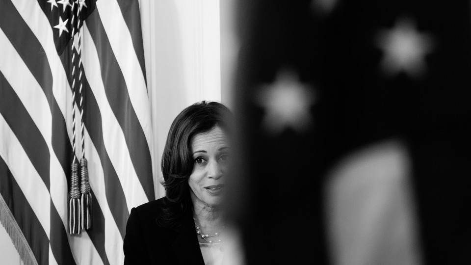 Vice President Kamala Harris is seen between two flags; one is blurred in the foreground and obscuring part of her face, and the other is just behind her.