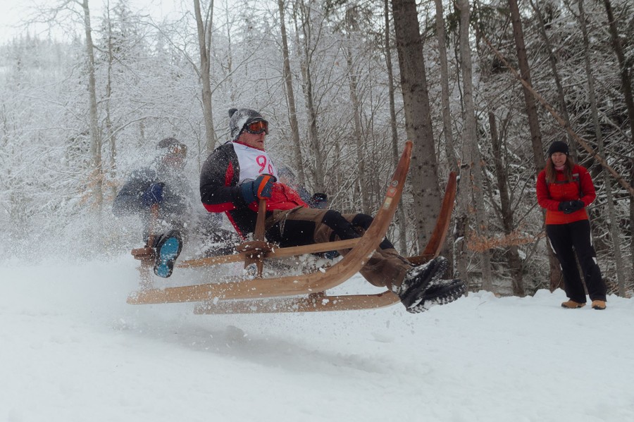 Two people are momentarily airborne as they ride a wooden sled down a snowy track.