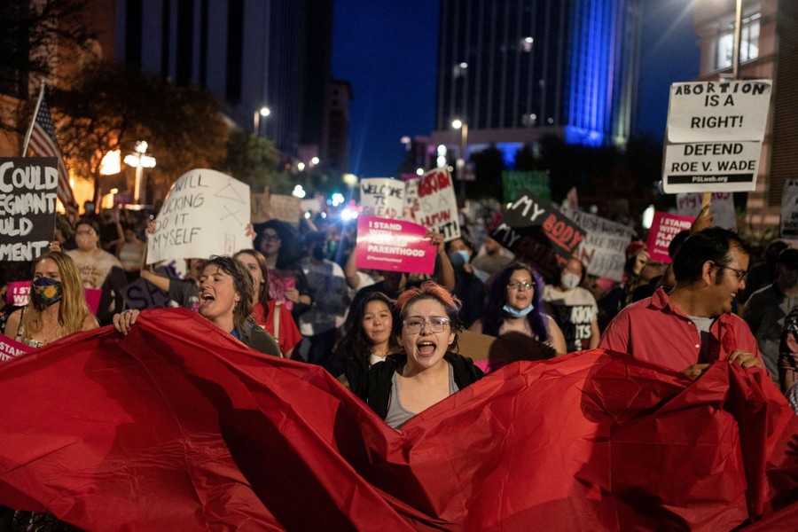 Dozens of protesters marching in a street, with those in front holding up a large red banner.