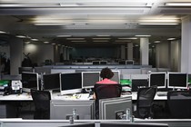 A worker sits alone in a dimly lit office, surrounded by computer monitors