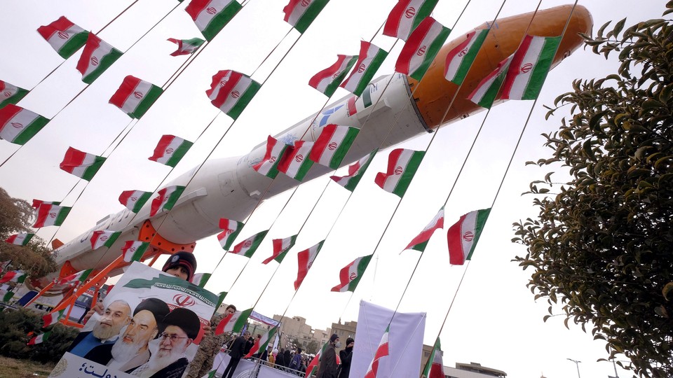 A boy holding a placard with pictures of President Hassan Rouhani, Ayatollah Ruhollah Khomeini, and Ayatollah Ali Khamenei, poses for camera in front of a model of Simorgh satellite-carrier rocket with Iranian flags everywhere. 