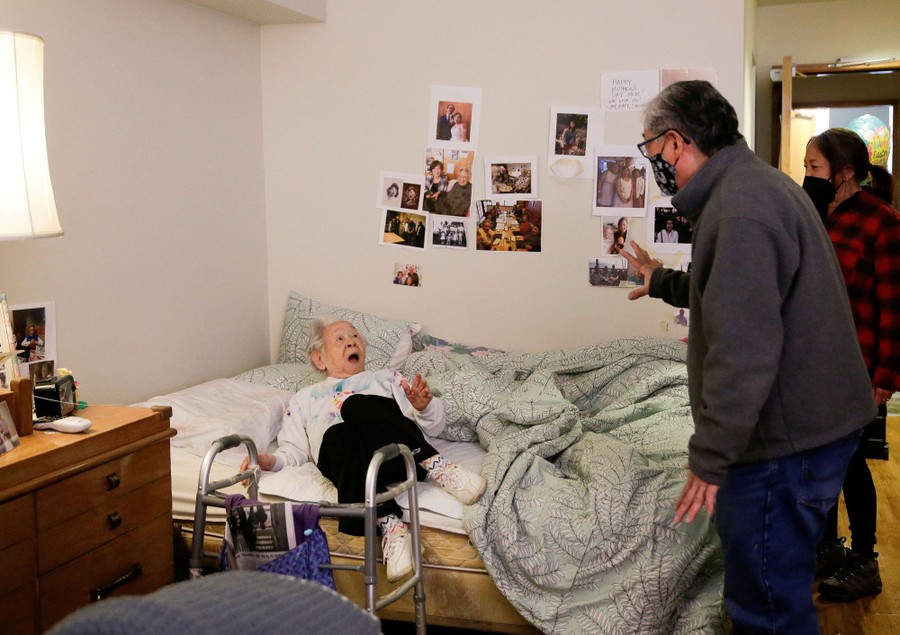 An older woman, lying on a bed, reacts with surprise as her son greets her.