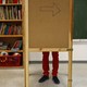 A pair of legs are visible behind a wooden corkboard in a classroom. 