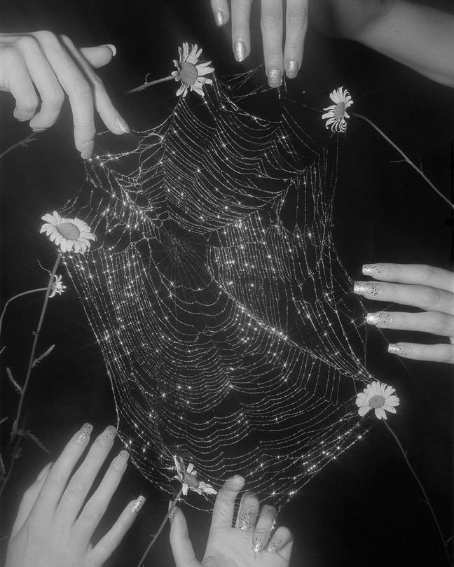 A black and white photograph of a large spider web surrounded by women's hands, all with long shiny nails