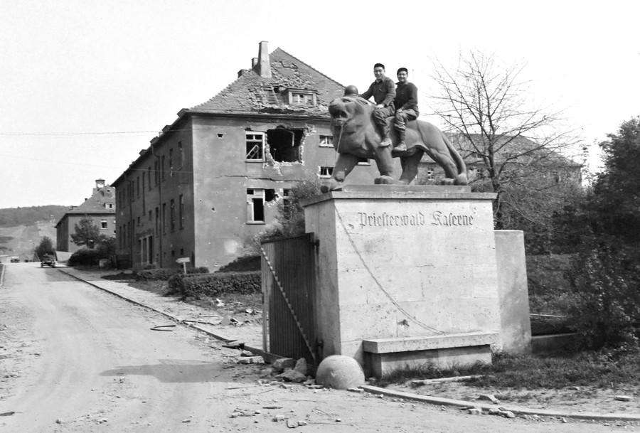 Two American soldiers sit on the back of a lion statue on a pedestal, in front of bomb-damaged military barracks.