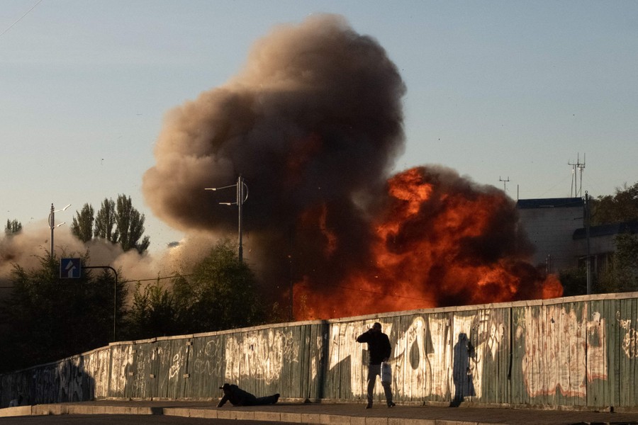 Two men are seen next to a fence one is laying on the ground, and on the other side of the fence a large fiery explosion rises into the air.