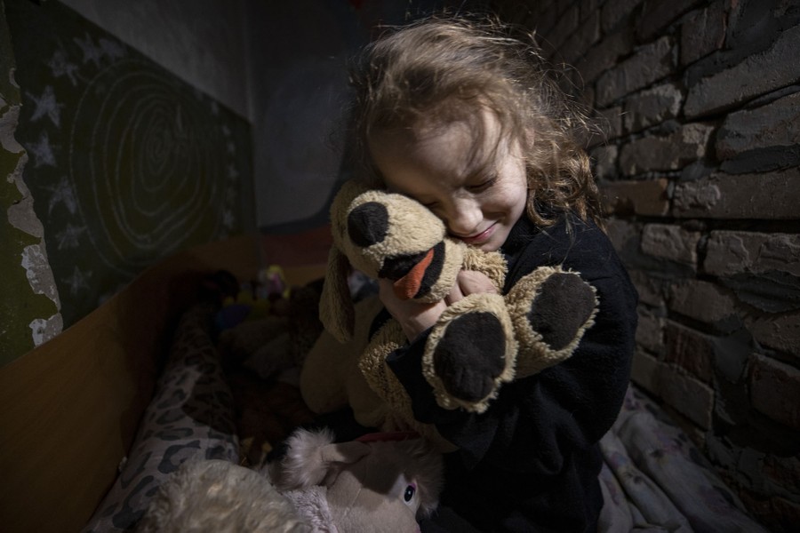 A girl squeezes a stuffed toy inside a brick-walled room.