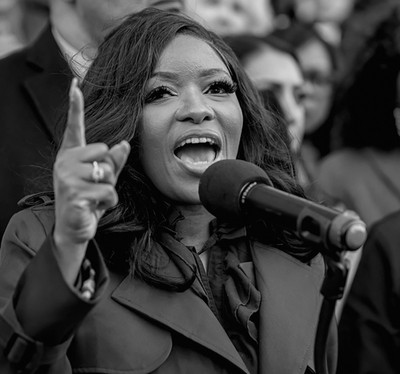 black-and-white photo of woman in suit speaking at mic and pointing upward with index finger