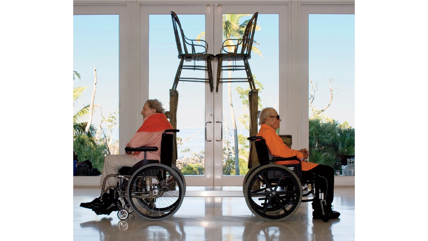 photo of two men in wheelchairs facing away from each other, each in front of the mirrored stair-step portions of Rauschenberg's 1981 sculpture, with the facing chairs visible at top and window overlooking palm trees and ocean behind