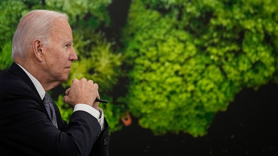 President Biden, in profile in front of a wall of green plants, gazes forward with his hands clasped.
