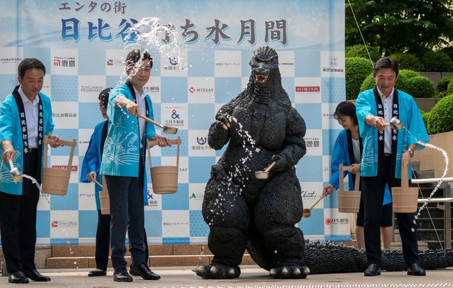 Several people, including one wearing a full-body Godzilla costume, use ladles to sprinkle water onto a stage.