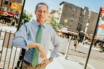 Color photo of Anthony Weiner standing near a busy urban intersection, his elbow resting on a fence.