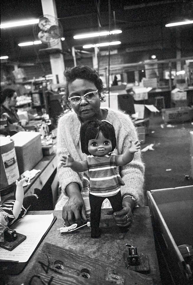 Black and white photo of factory floor with Black woman in cat-eye glasses holding a standing Remco doll