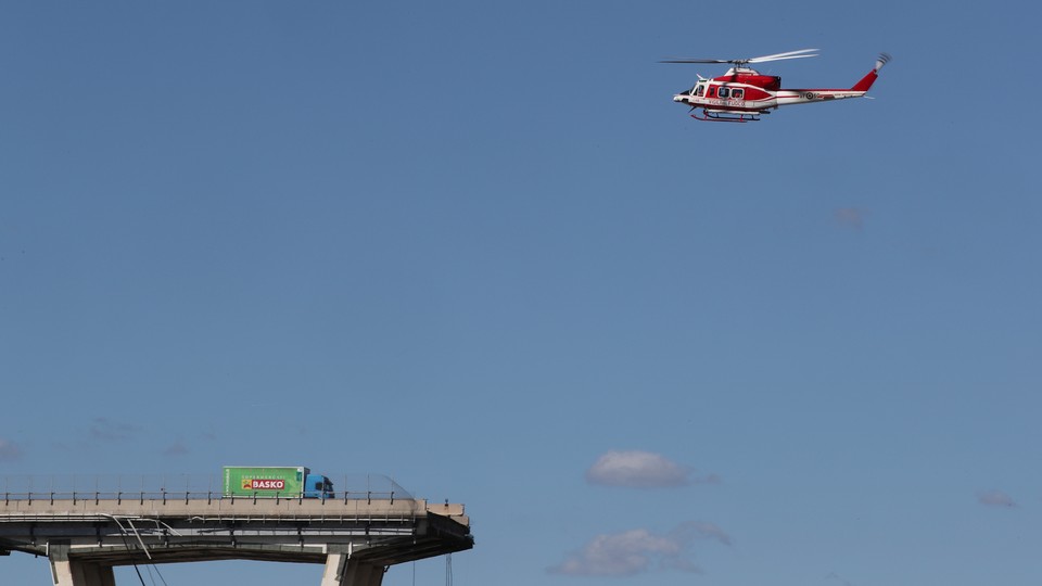 A firefighter helicopter flies above the destroyed Morandi bridge, where a truck pauses just before the section that broke apart.