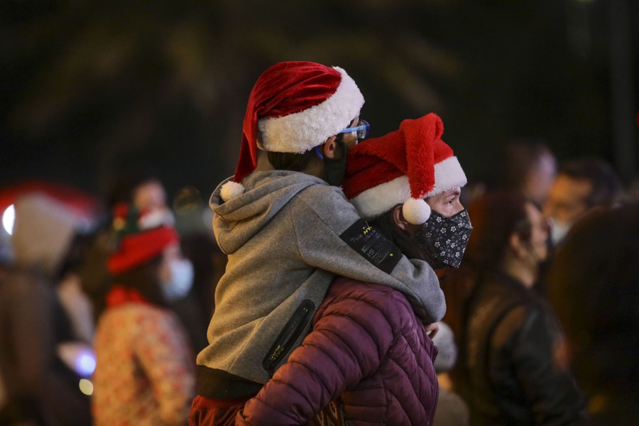 A person carries a child on their shoulders in a crowd, both wearing Santa hats.