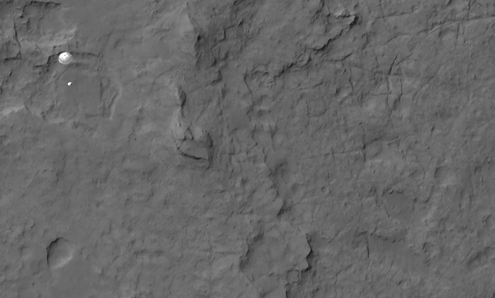 A high orbital view of the surface of Mars, with a small parachute and space capsule visible at upper left.