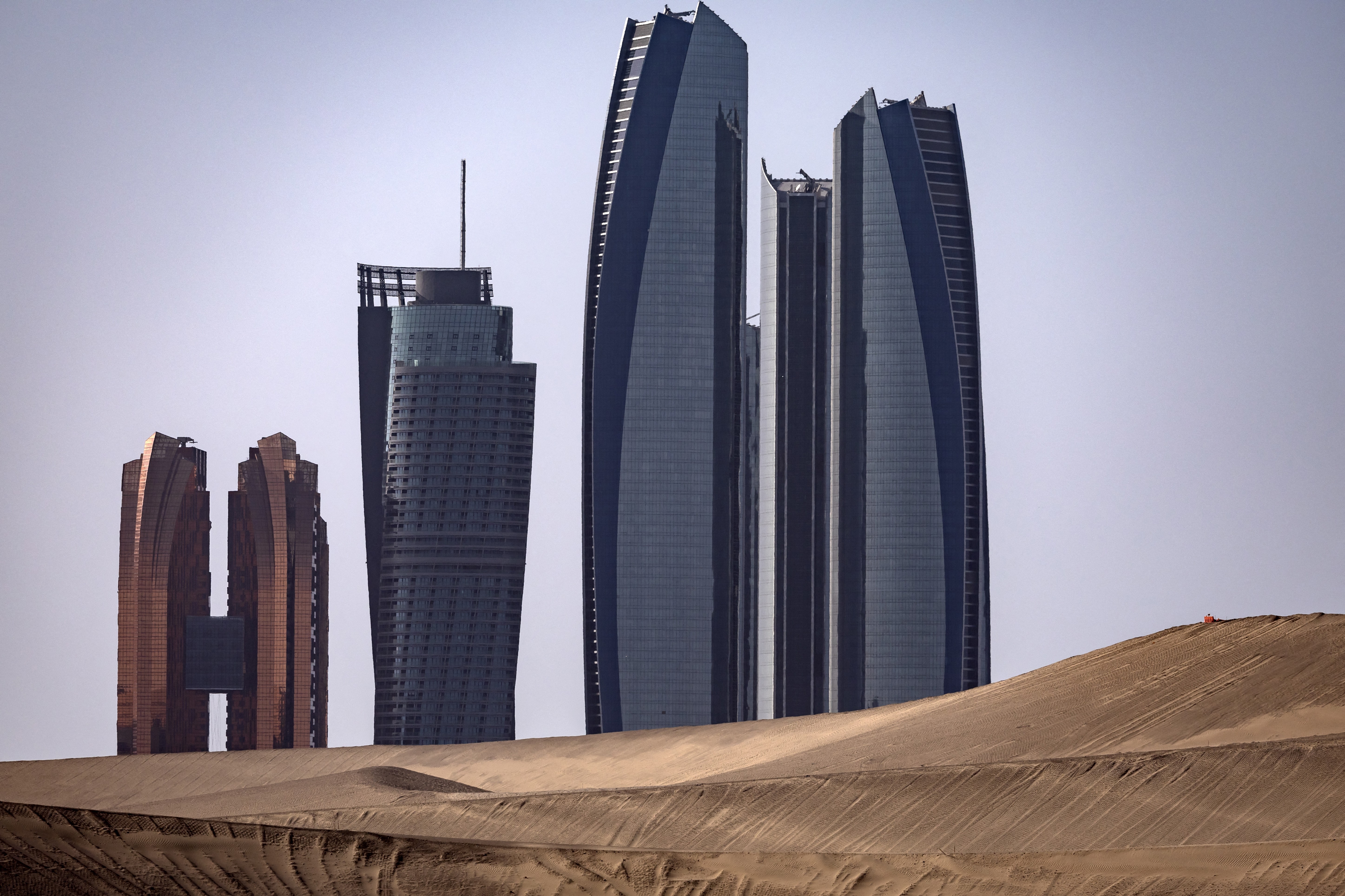 A cluster of modern skyscrapers, seen past sand dunes.
