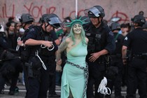 Police officers in riot gear arrest a protester dressed as Lady Liberty in chains.