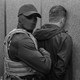 a black and white photo of a masked ICE agent detaining a man in immigration court.