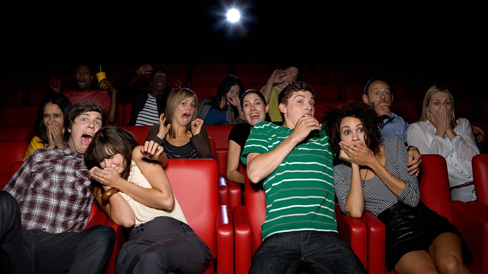 A group of people in a movie theater make scared faces and clutch each other.