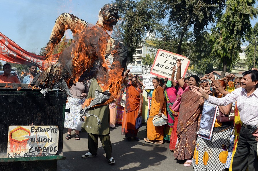 Several people are gathered at a protest. An effigy is burning, and signs read "Extradite Union Carbide" and "Killer Carbide is now Dow Chemical."