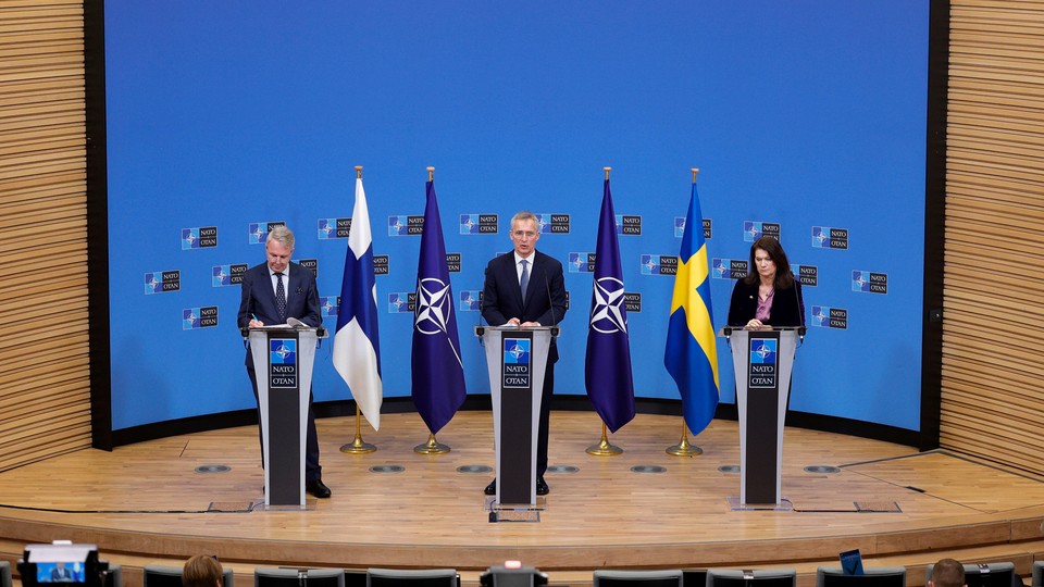 Three people stand at lecterns on a stage, with the flags of Finland, Ukraine, and NATO between them, during a press conference in Brussels.