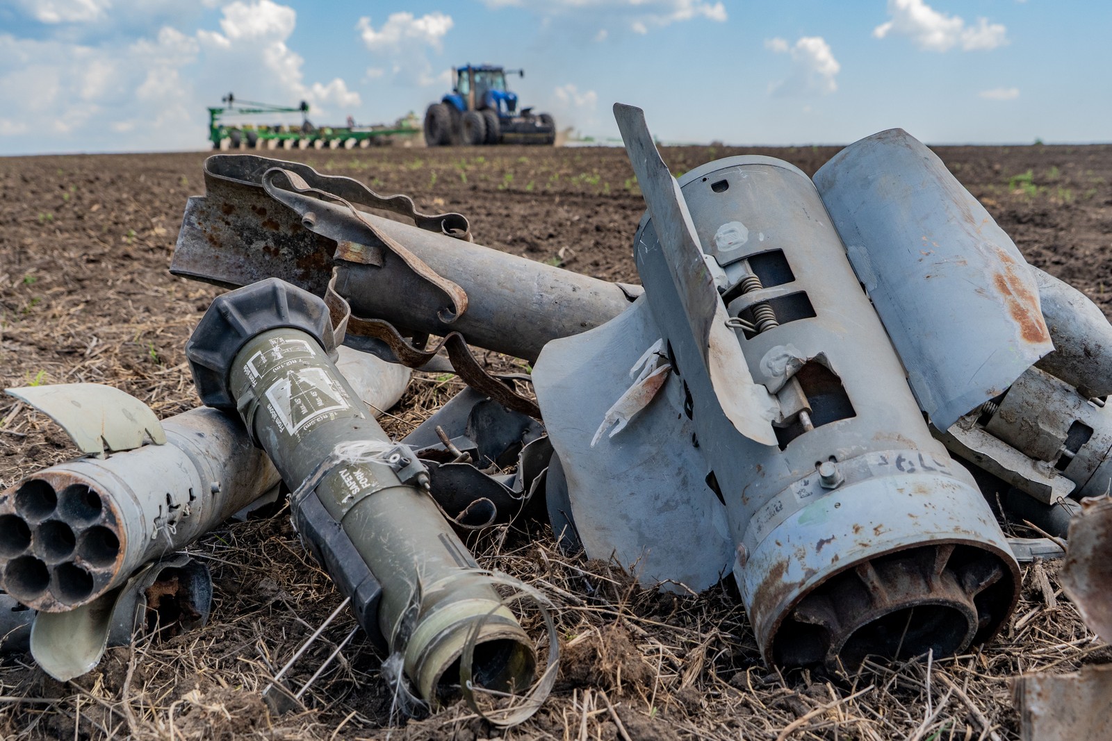 Fragments of Russian missiles and weapons lie piled on a field as a tractor passes by in the background.