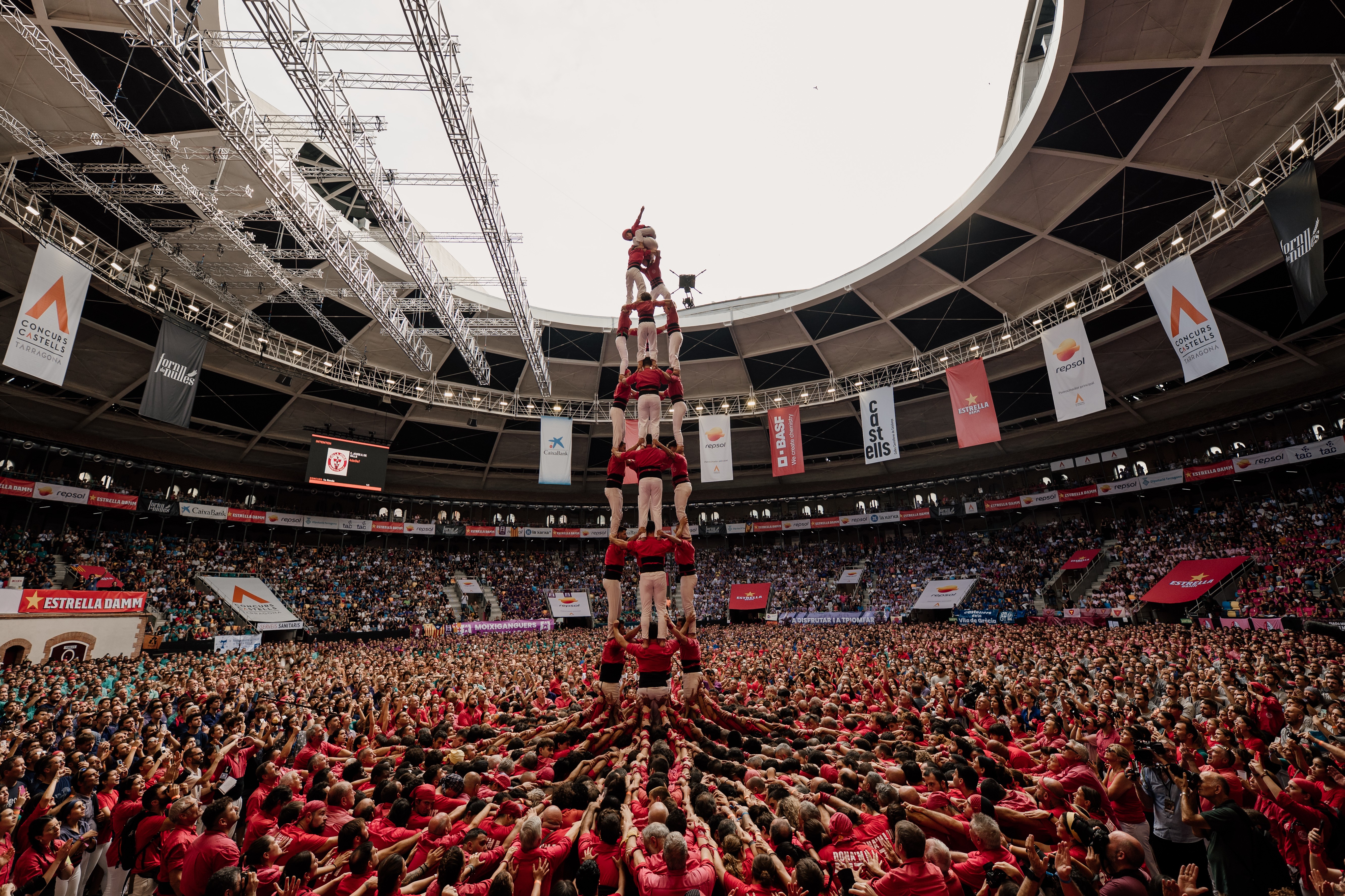 A crowd in an arena watches as a team forms a human tower, surrounded by other teams.