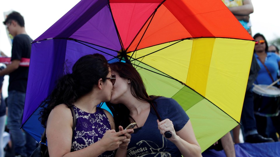 Two women kiss under a rainbow umbrella.