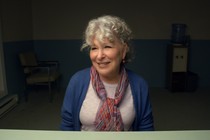 The actor Bette Midler sits at a desk wearing a white shirt and a blue sweater