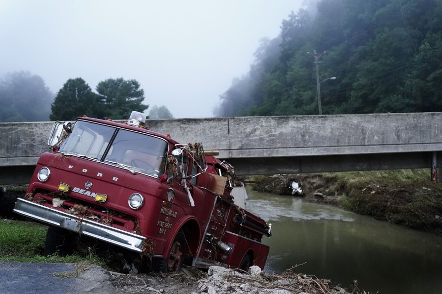 A fire truck sits at an angle, jammed up beneath a concrete bridge along a river bank.