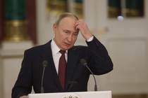 Vladimir Putin stands at a lectern, looking downward and holding his hand up to his head.