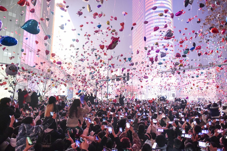 A crowd in a brightly lit city square releases hundreds of balloons into the air.