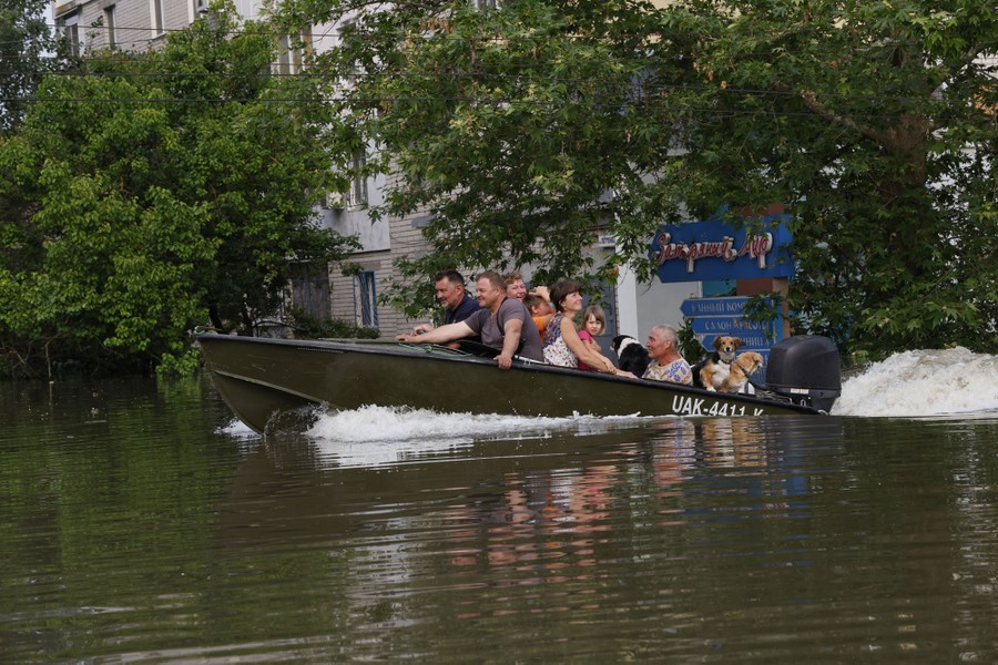 A small boat carries seven or eight people and several pet dogs through a flooded area.