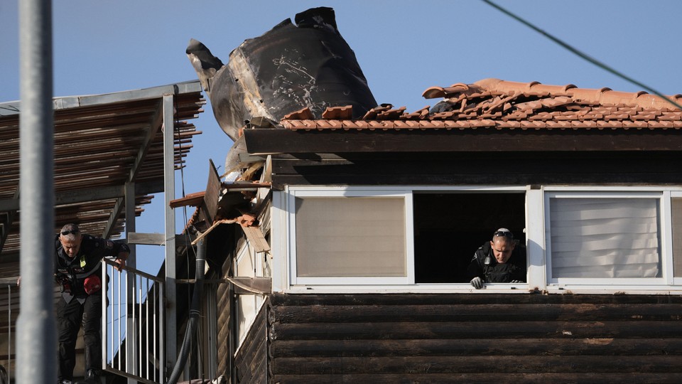 A color photograph of a man peering out the window of a house whose roof appears to have been destroyed by an explosion.