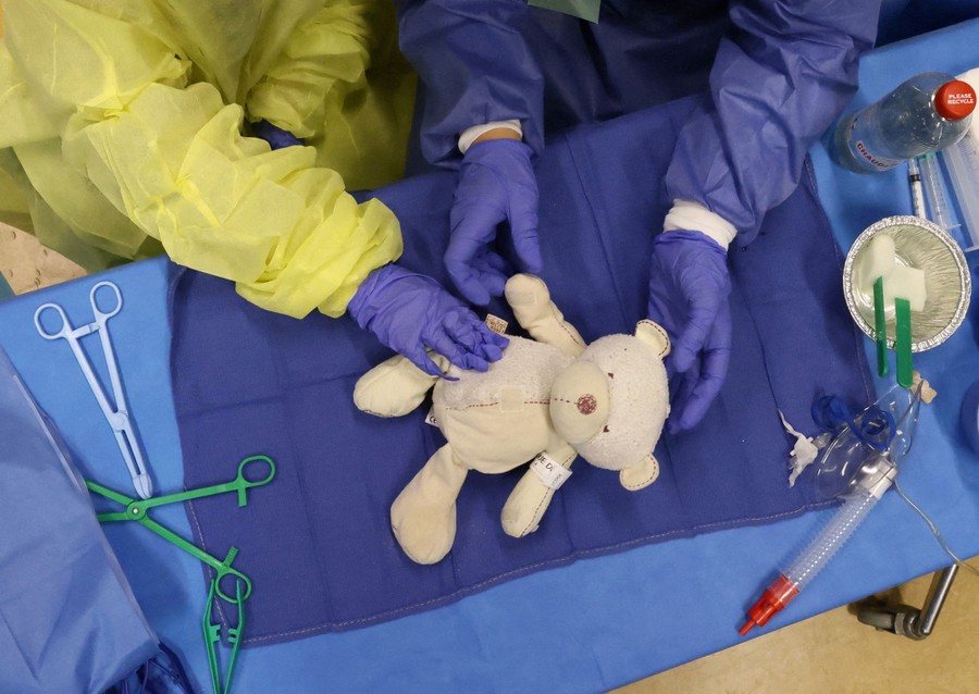A view looking down on a teddy bear on a table being attended to by two people wearing surgical gloves and protective gear