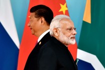 Xi Jinping and Narendra Modi stand next to each other but face in opposite directions in front of flags.