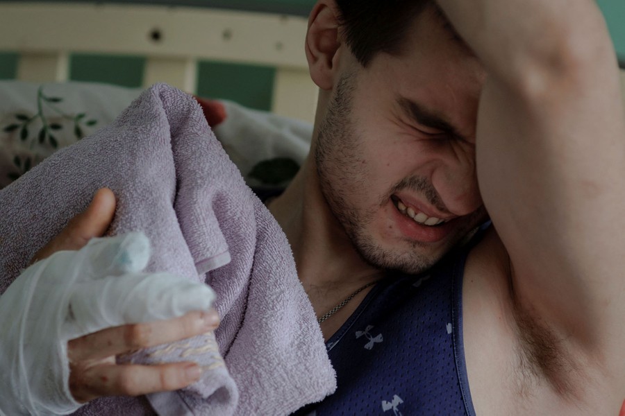 A man grimaces while holding one arm over his head, the other (bandaged) in front of him, in a hospital bed.