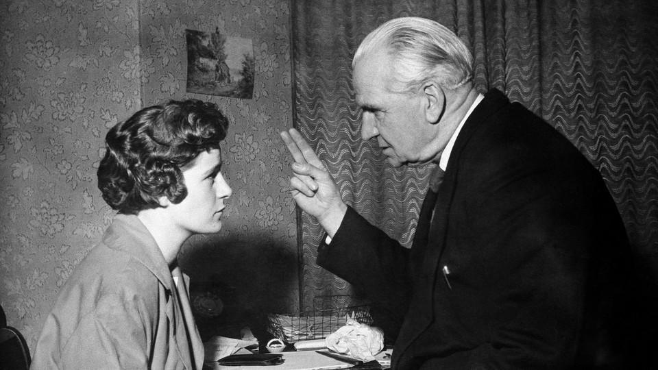 In a black-and-white photo, an old man holds his fingers in front of a young girl's face.