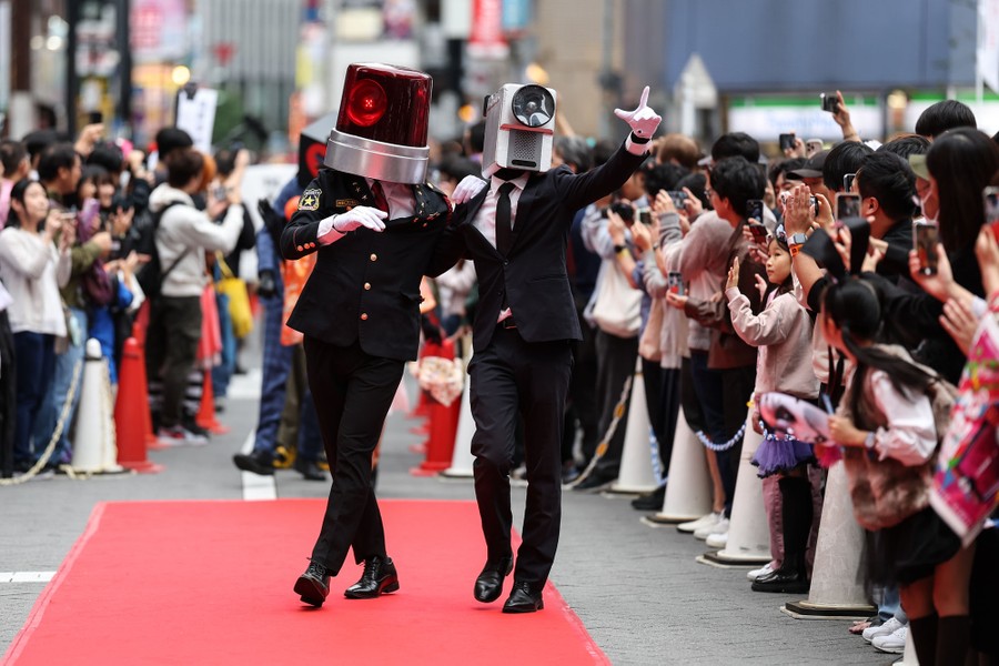 Two people in costume, wearing full-head masks of a police siren and a video camera, walk along a red carpet, past a crowd.