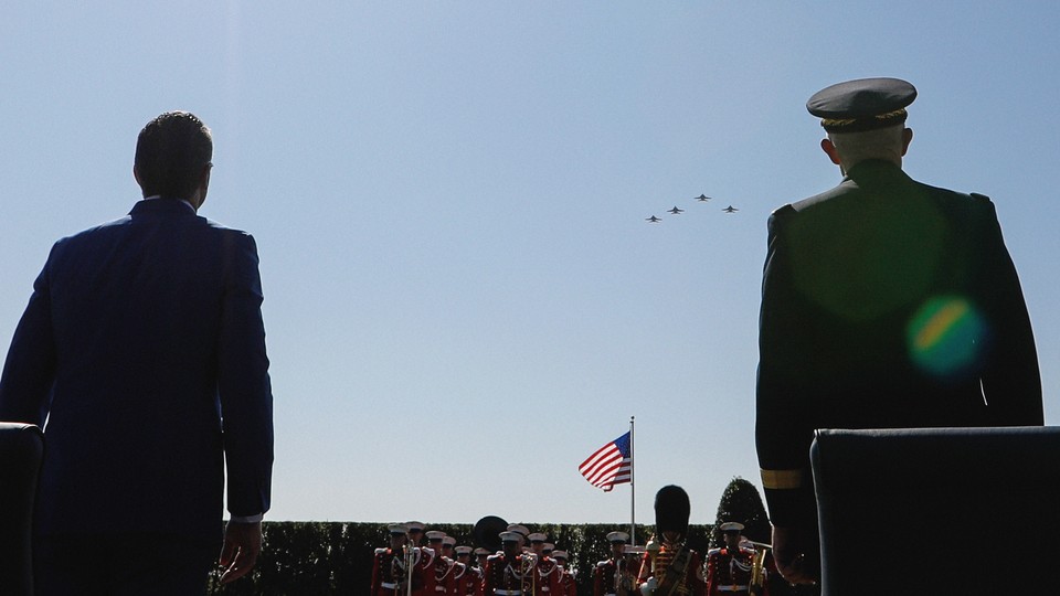 Photograph of two figures (left, Pete Hegseth) seen from behind, looking up at a fighter jet formation flying in a blue sky