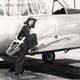 black-and-white archival photo of woman in flight gear and cap, smiling at camera, standing with one foot on ground and one foot on the wing of the plane she piloted, about to climb into cockpit