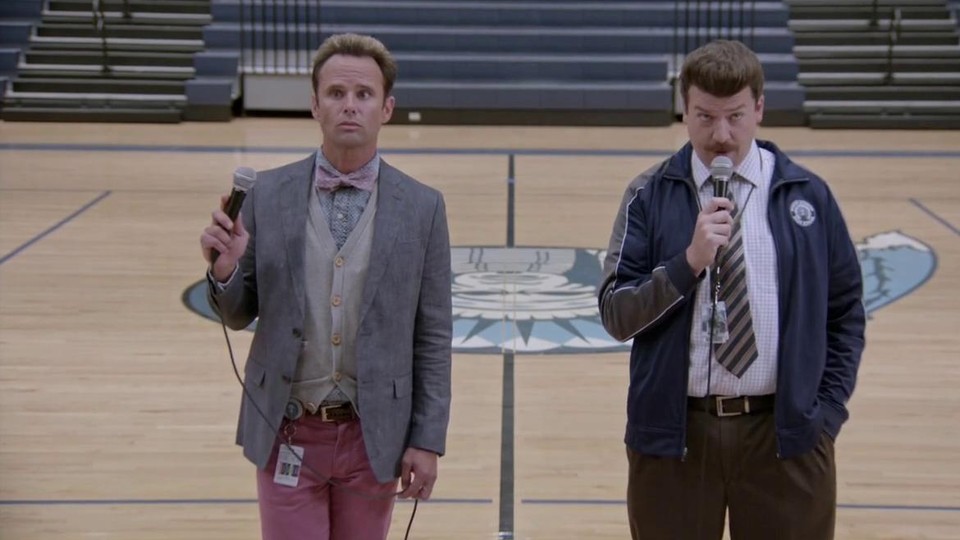 Walton Goggins, as Lee Russell, and Danny McBride, as Neal Gamby, stand in the center of a gymnasium holding microphones. 