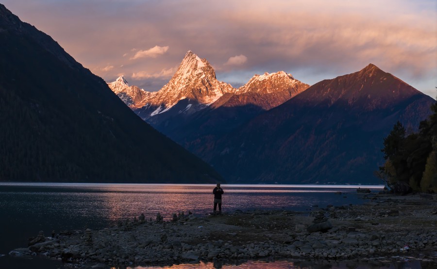 A person stands beside a lake, looking toward distant steep mountains.