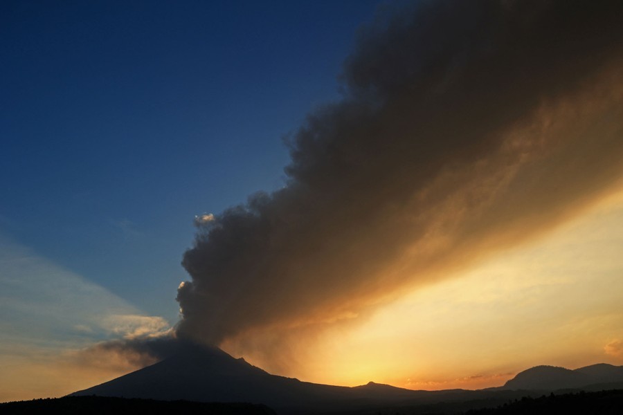 A volcano spews ash into the sky, casting shadows against a low sun.