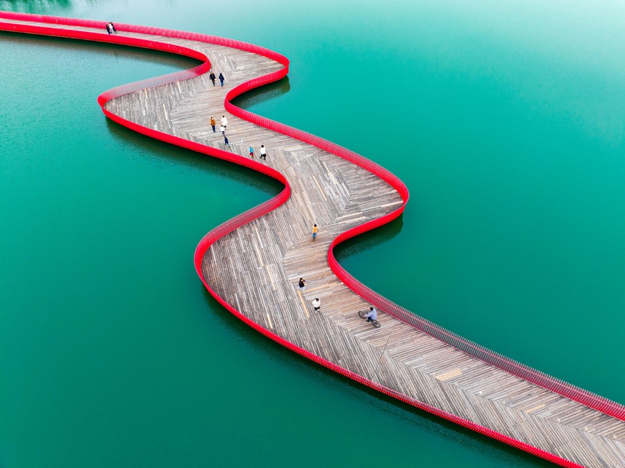 A wooden walkway lined in red snakes across a teal-colored lake.