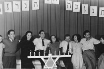 In a black and white photo, a group of men and women laugh and smile in a row standing behind a menorah.