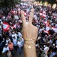 A close up photo of a demonstrator flashing a V sign. In the background, protesters gather waving Lebanon flags.