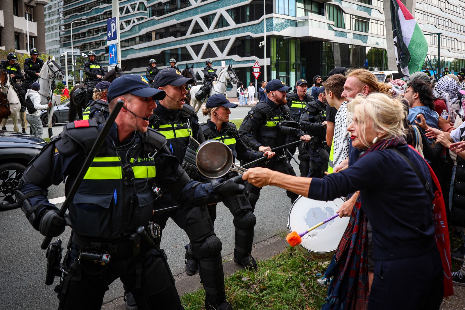 Police in riot gear face off against protesters, one of whom holds up a cooking pot, gesturing toward the police.