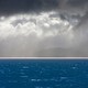 An image of storm clouds with sun shining through onto a hyper-blue ocean